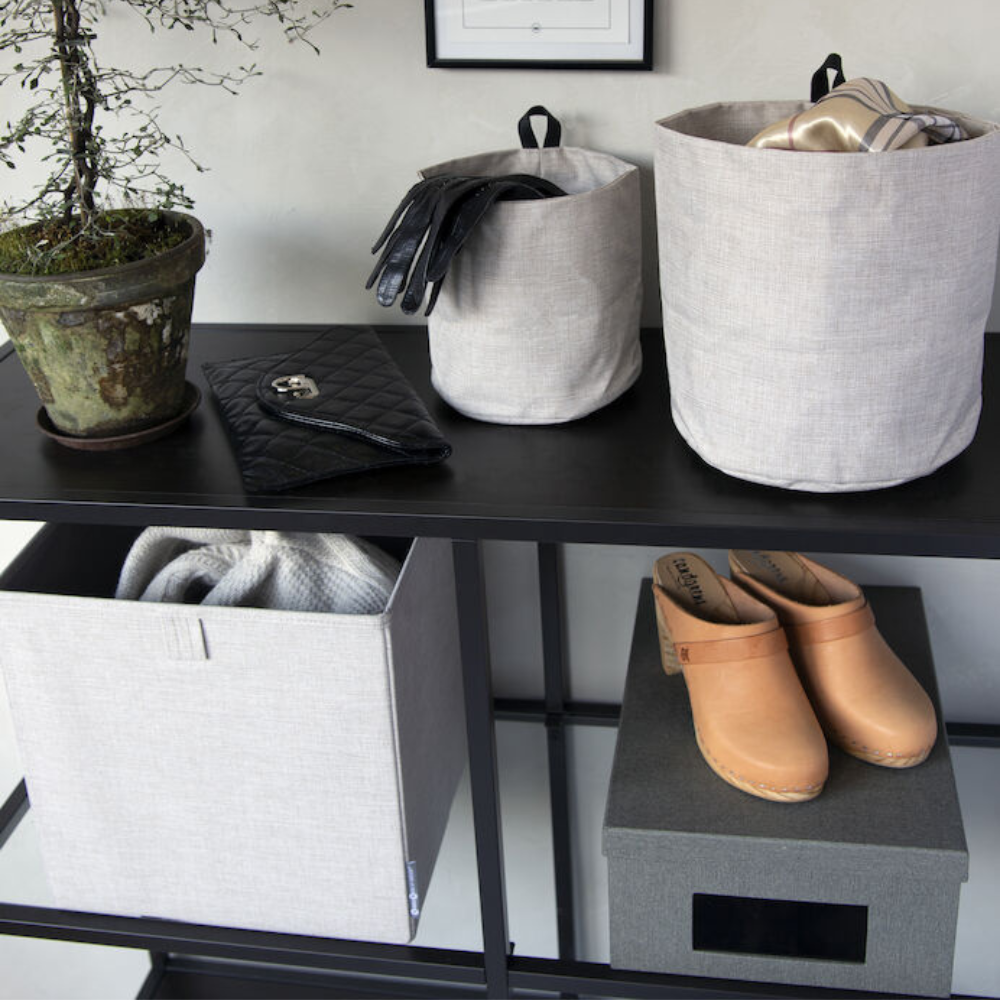 Ein schwarzes Regal enthält eine Topfpflanze, eine schwarze Handtasche, zwei Bigso SOFT Aufbewahrungsbox CUBE - Beige für Handschuhe und Schals, hellbraune Clogs und eine dunkle Aufbewahrungsbox. Neutrale Farbtöne der beigen Boxen sorgen für einen aufgeräumten, organisierten Look.