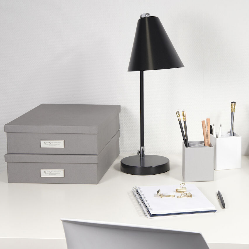 Ein aufgeräumter Schreibtisch mit zwei gestapelten Bigso OSKAR Aufbewahrungsboxen A4 in Grau, einer schwarzen Schreibtischlampe, einem Notizbuch mit goldenen Büroklammern und Behältern für Bleistifte und Kugelschreiber, alles vor einer weißen Wand.