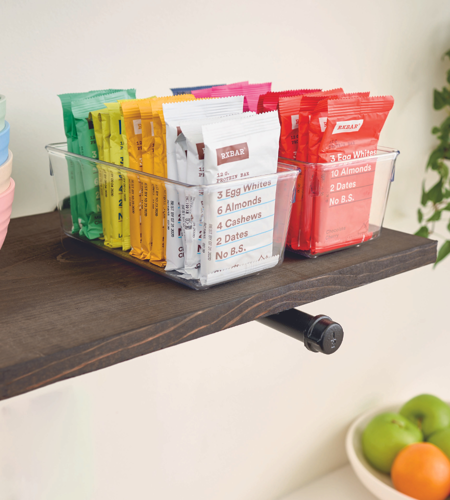 Un organizer per cassetti iDesign LINUS ALTO tiene in ordine le barrette proteiche colorate RXBAR su uno scaffale di legno scuro, sotto c'è una ciotola con della frutta.