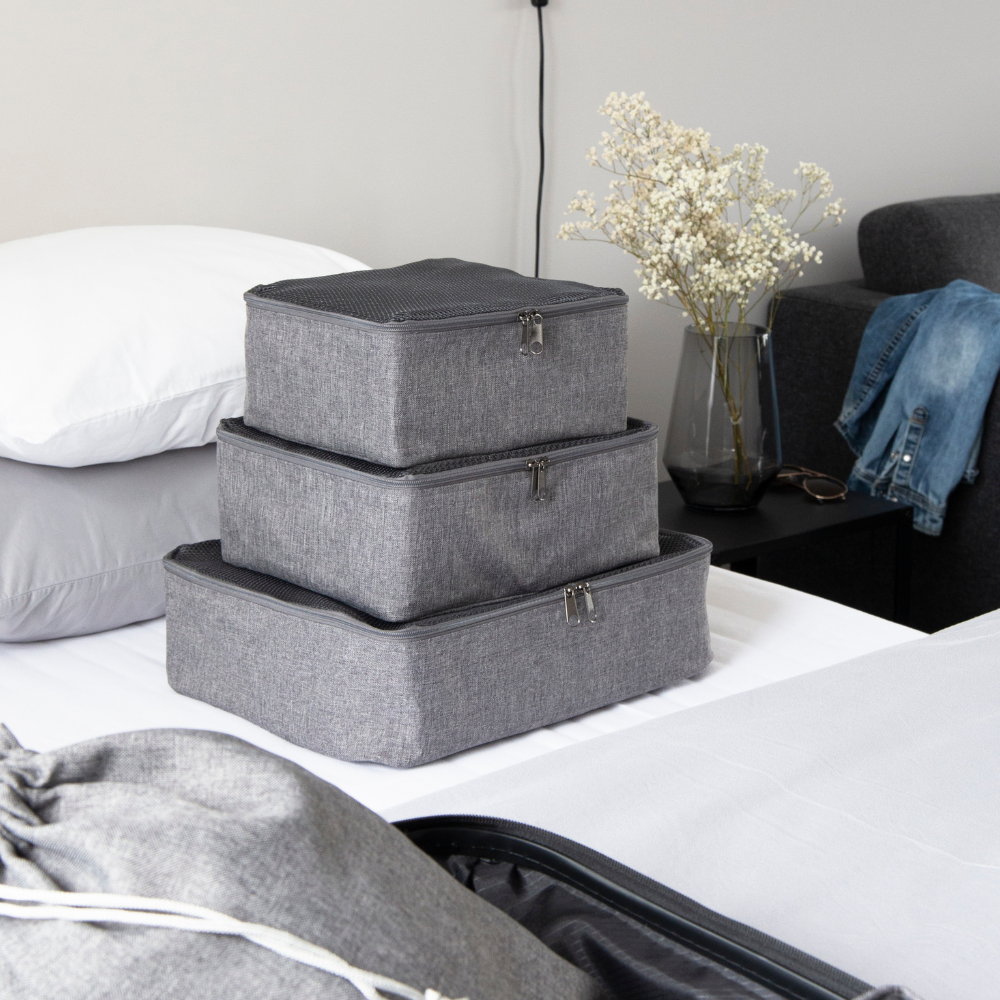 Drei graue Bigso TRAVEL Organizer M sind auf einem ordentlich gemachten Bett neben einem offenen Koffer gestapelt. Ein Nachttisch mit Trockenblumen, einer Brille und einem gefalteten Hemd vervollständigt die perfekte Umgebung für diese vielseitigen Utensilien.
