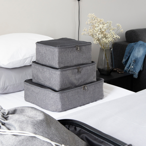 Drei graue Bigso TRAVEL Organizer M sind auf einem ordentlich gemachten Bett neben einem offenen Koffer gestapelt. Ein Nachttisch mit Trockenblumen, einer Brille und einem gefalteten Hemd vervollständigt die perfekte Umgebung für diese vielseitigen Utensilien.