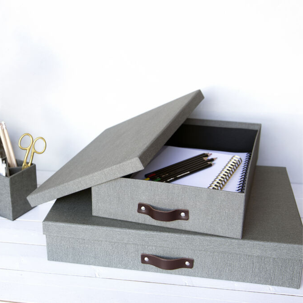 Eine graue Bigso SVERKER Aufbewahrungsbox A3 aus Canvas mit einem Griff liegt geöffnet auf einer weißen Fläche und zeigt Notizbücher und Stifte im Inneren. Darunter befindet sich eine weitere geschlossene Box mit einem kleinen Behälter mit Stiften und einer Schere in der Nähe.