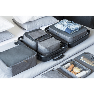 Ein ordentlich organisierter offener Koffer mit dem TRAVEL Organizer L - Grau von Bigso und Taschen sowie Kleidung ist umgeben von Toilettenartikeln in Reisegröße, einem Kamm und einer Bürste - alles auf einem Bett mit weißen Kissen im Hintergrund.