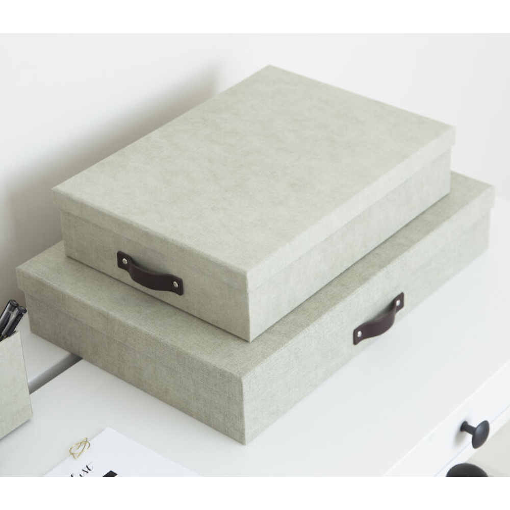 Zwei gestapelte hellgraue Bigso OSKAR Aufbewahrungsboxen A4 - Linnen Canvas mit schwarzen Griffen stehen auf einer weißen Fläche, ideal für die Aufbewahrung von Dokumenten in Ihrem Heimbüro. In der Nähe befinden sich ein kleines Tablett und einige Papierartikel.