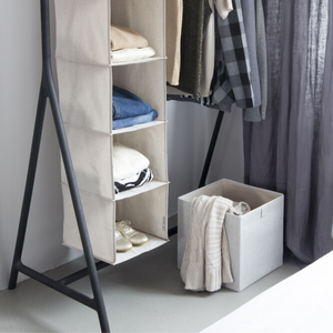 Ein Hängeorganizer aus Stoff mit Einlegeböden nimmt Kleidung und Schuhe auf. Daneben steht eine Bigso SOFT Aufbewahrungsbox CUBE in Beige, die einen gefalteten Pullover enthält, während mehrere Hemden und Jacken auf einem schwarzen Kleiderständer daneben hängen.