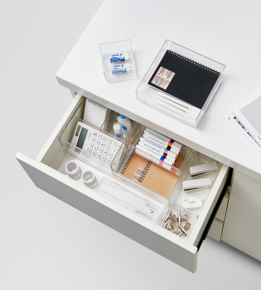 An open desk drawer holds the BNB x VCN SLIDE drawer organizer KLAR - various sizes, which keeps pens, highlighters, calculators, tape, erasers, notebooks, and paper clips neatly sorted; on top are a closed notebook and erasers. Perfect for organizing drawers.