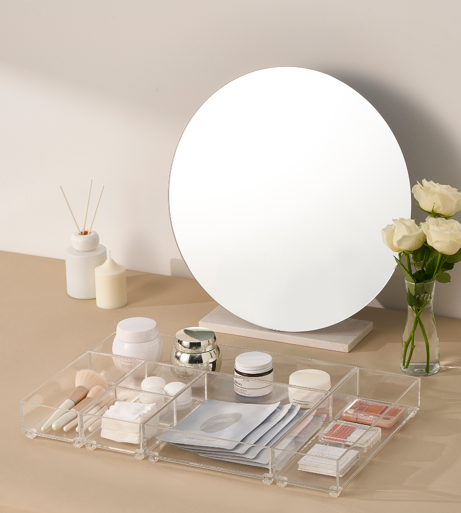 Uno specchio rotondo è appoggiato su un lavabo beige con rose bianche e candele. Pennelli per il trucco, creme, prodotti per la cura della pelle e maschere in tessuto sono sistemati in un organizer e nel set di cassetti SLIDE (5 pezzi) di BNB x VCN.