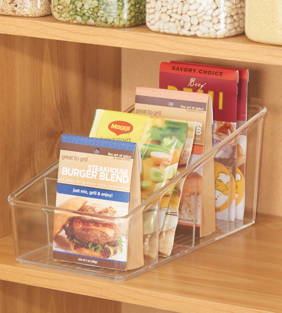 Der iDesign LINUS Aufbewahrungsbehälter KLAR mit Fächern bringt Ordnung in ein hölzernes Vorratsregal für Gewürz- und Saucenpackungen, während andere Trockengutbehälter über ihm aufbewahrt werden.
