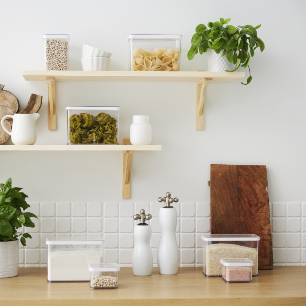 Kitchen shelf with storage containers for pasta and legumes, surrounded by plants and dishes.