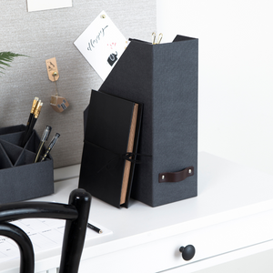 Eine Bigso ESTELLE Stehsammler Büroablage in Schwarz Canvas und ein Notizbuch liegen auf einem weißen Schreibtisch neben einem Stifteköcher mit schwarzen Stiften. Im Hintergrund befindet sich eine Korkplatte mit Notizen, und im Vordergrund ist ein Teil eines schwarzen Stuhls zu sehen.