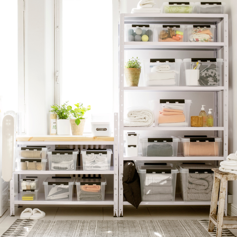 Offene Regale zeigen die Orthex SmartStore Box KLAR 40x30x11cm (7,5L) Flach, BPA-frei und beschriftet, neben gefalteten Handtüchern, Toilettenartikeln und Topfpflanzen in einem hellen Raum mit einem gemusterten Teppich.