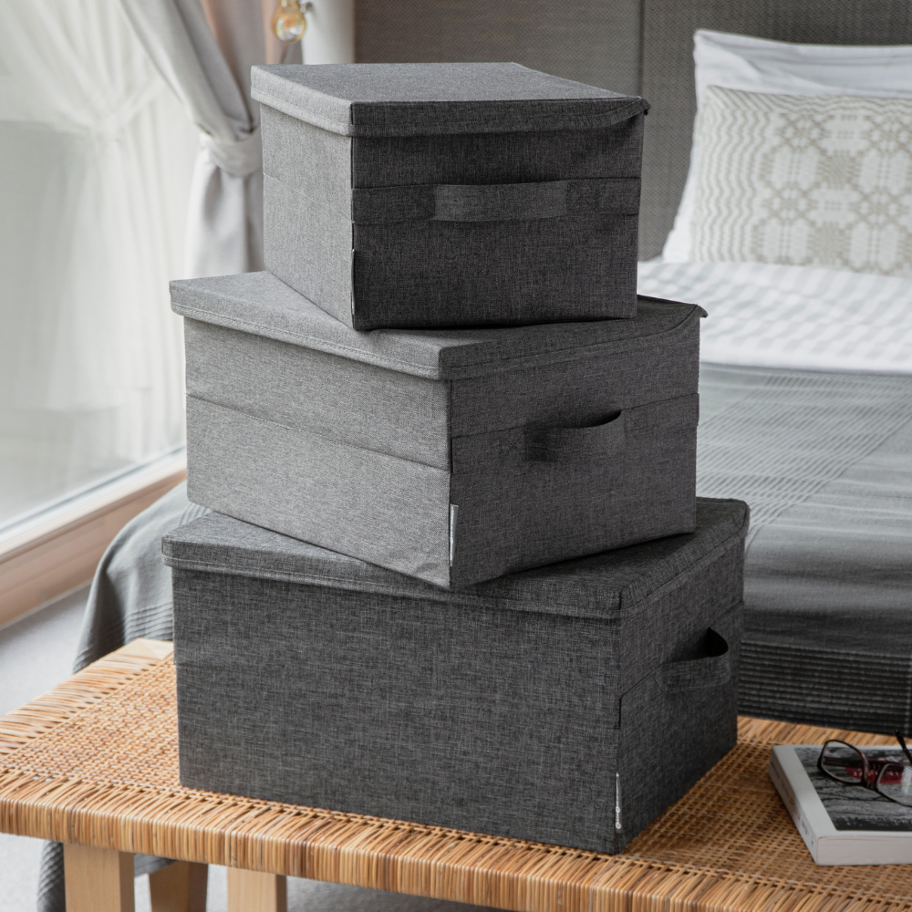 Drei Bigso SOFT Aufbewahrungsboxen Grau M (40x30x22cm) sind im Schlafzimmer auf einer geflochtenen Bank gestapelt. Im Hintergrund befindet sich ein ordentlich gemachtes Bett und ein großes Fenster.