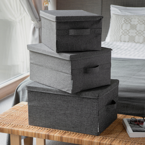 Drei Bigso SOFT Aufbewahrungsboxen Grau M (40x30x22cm) sind im Schlafzimmer auf einer geflochtenen Bank gestapelt. Im Hintergrund befindet sich ein ordentlich gemachtes Bett und ein großes Fenster.