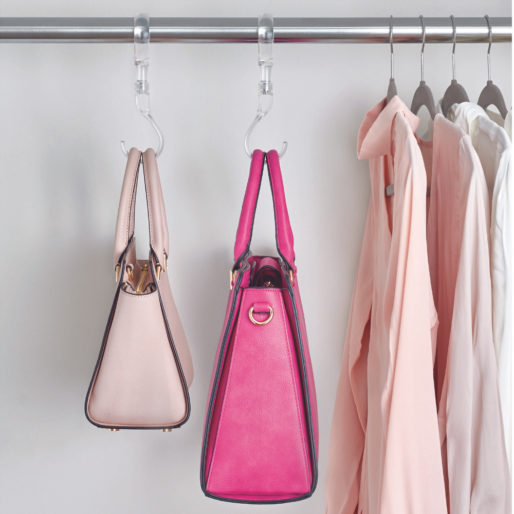 Zwei Handtaschen, eine hellrosa und eine knallrosa, hängen platzsparend von iDesign HOME EDIT - S-Haken KLAR an einer Metallstange neben mehreren hellen Hemden auf Bügeln vor einer weißen Wand.