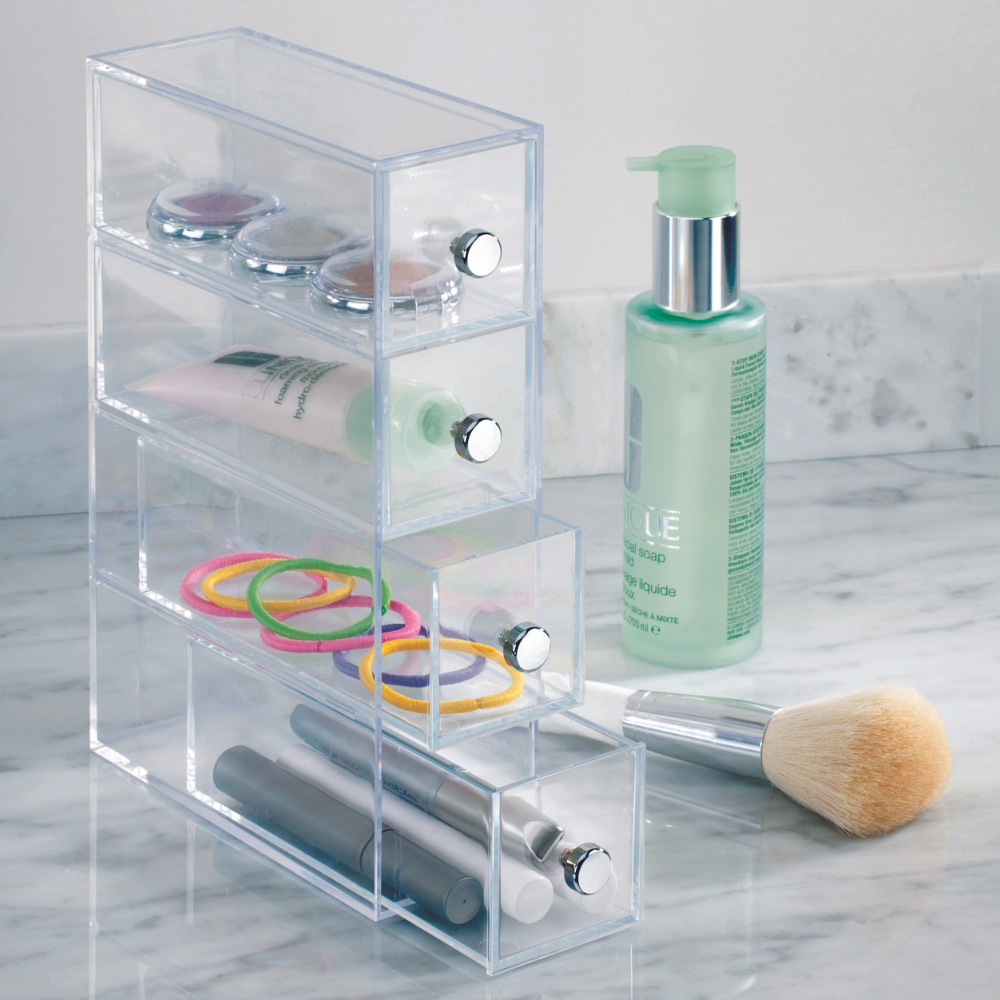 Der iDesign DRAWER - Schubladen-Turm KLAR 4 Fächer - Schmal steht auf einer Marmortheke und bewahrt Schminke, Haargummis und Pinsel ordentlich auf. Daneben stehen ein Schminkpinsel und eine grüne Pumpflasche mit Gesichtsseife oder -lotion.