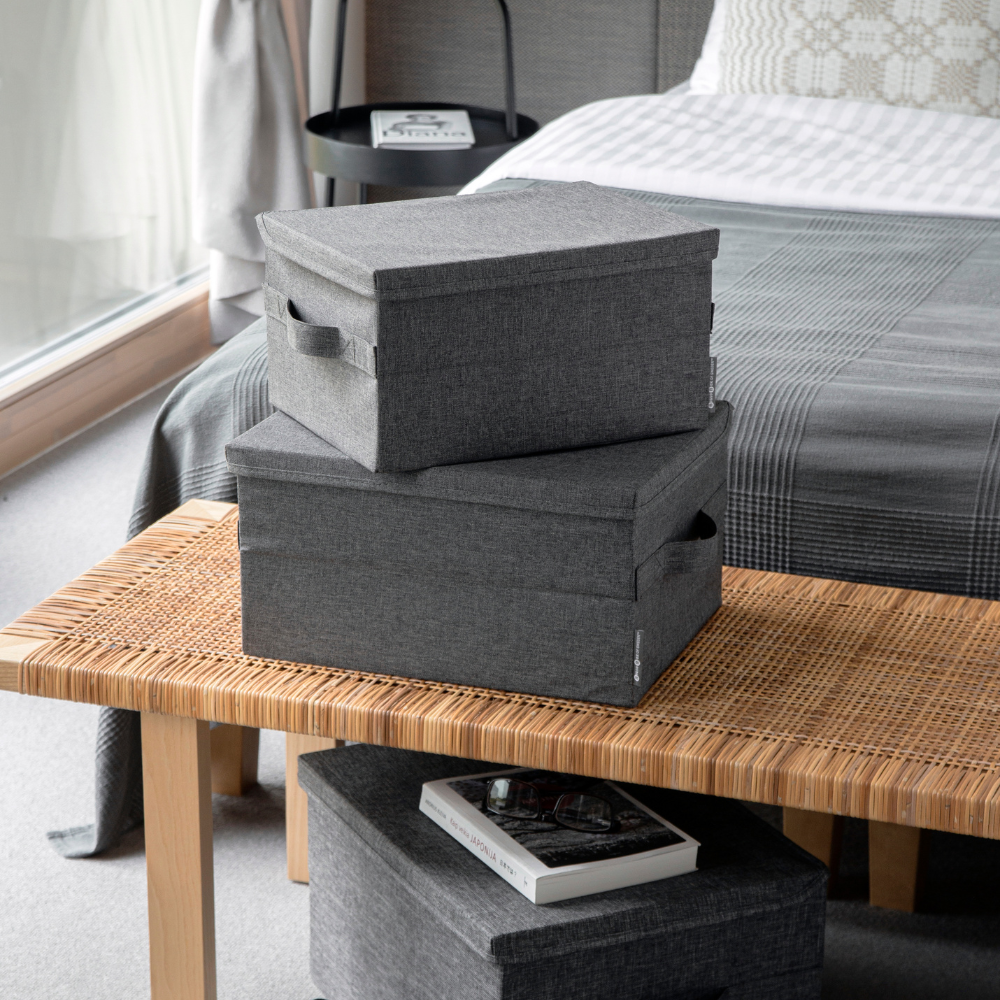 Zwei SOFT Aufbewahrungsboxen Grau L 45x34x25cm von Bigso sind auf einer geflochtenen Bank am Fußende eines Bettes gestapelt. Eine kleinere Box und ein Buch liegen daneben, alles sanft beleuchtet vom natürlichen Licht des Fensters.