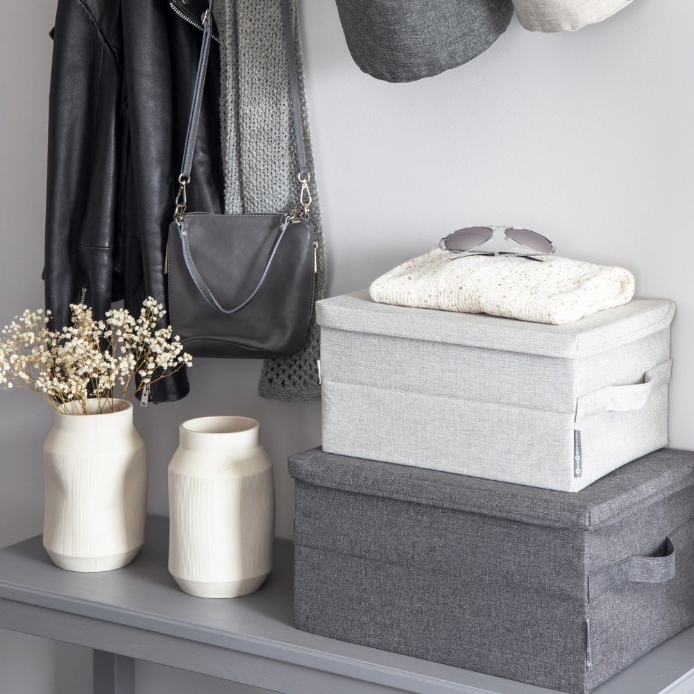 Ein grauer Tisch hält zwei Bigso SOFT Aufbewahrungsbox Beige S (35x26x19cm). Obenauf liegen gefaltete Kleidung und eine Sonnenbrille. Zwei weiße Vasen mit getrockneten Blumen stehen in der Nähe, während eine Lederjacke und eine Handtasche darüber hängen.