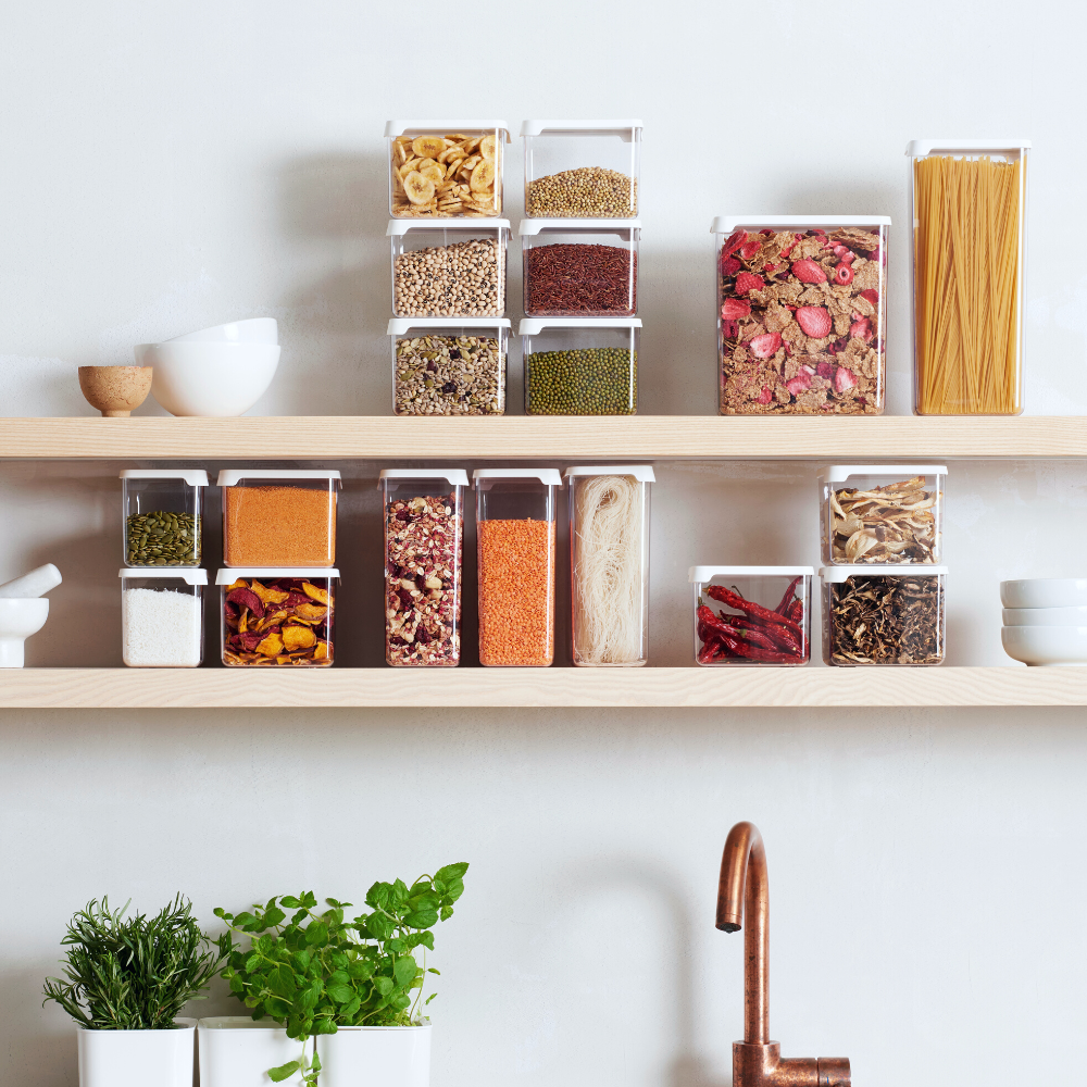 I contenitori per alimenti SmartStore TRASPARENTI - Bianchi di Orthex, pieni di cereali, pasta e spezie, sono disposti ordinatamente su due scaffali in legno; in basso ci sono dei vasi con le erbe aromatiche, e sotto c'è un rubinetto in rame.