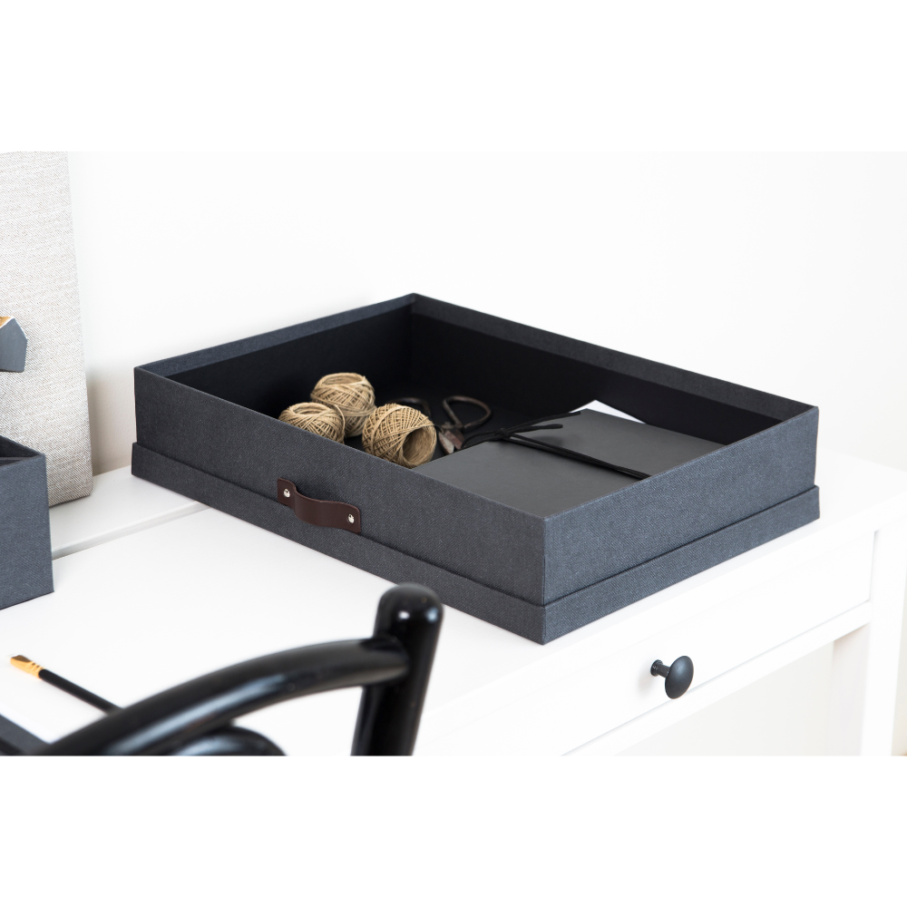 Eine SVERKER Aufbewahrungsbox A3 in Schwarz Canvas von Bigso steht auf einem weißen Schreibtisch, auf dem Garnrollen, Scheren und Notizbücher ordentlich aufbewahrt werden. Ein schwarzer Stuhl und ein Bleistift vervollständigen die minimalistische, organisierte Szene.