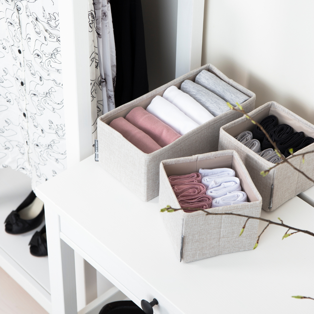 Kleine Stoffboxen mit gefalteter Kleidung auf weißem Tisch, daneben Kleiderschrank und Ast mit Knospen.